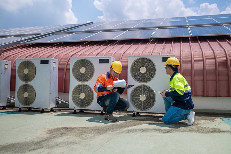 Two technicians in hard hats inspecting outdoor HVAC units on a rooftop below solar panel.