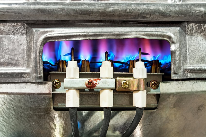 A close-up view of active blue gas flames burning inside a furnace or water heater burner assembly.