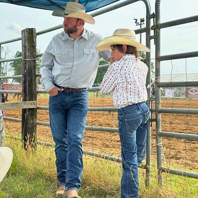 Electrical manager Johnny B and his son, both in cowboy boots and cowboy hats, leaning against a fence at a rodeo event.