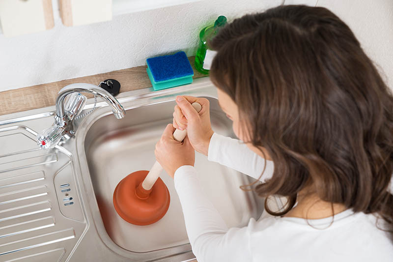 Woman Using Plunger In Kitchen Sink At Home, Garbage Disposal
