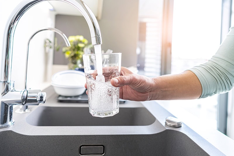 A person filling a glass with fresh tap water from a modern kitchen faucet over a sink.