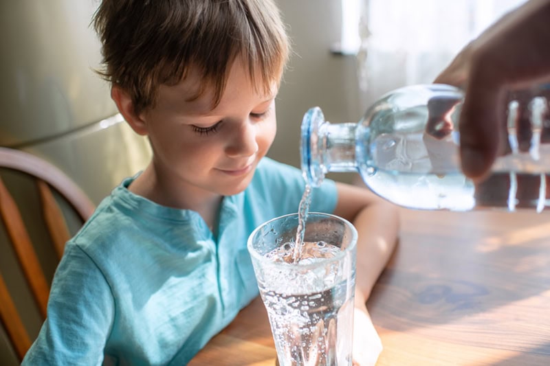 A young boy sitting at a table smiling as water is poured from a glass bottle into a drinking glass.