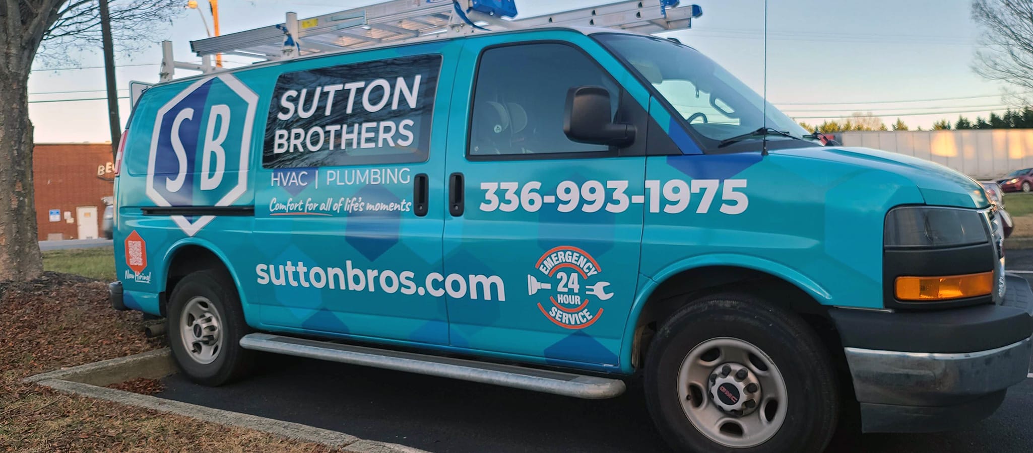 A Sutton Brothers GMC Savana truck branded with a teal hexagonal pattern and a ladder rack on the roof parked outside.